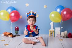 a baby sitting on the floor wearing a birthday hat and holding a toy in front of balloons