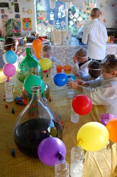 several children are sitting at a table with balloons and water bottles on it, while adults stand in the background