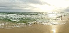 a person standing in the surf on a beach with waves coming towards shore and sun shining through clouds