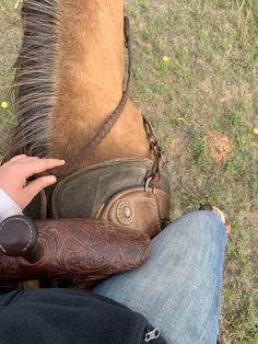 a person sitting next to a brown horse on top of a grass covered field with a saddle