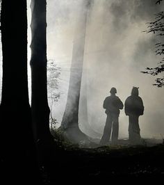 two people standing next to each other in the woods on a foggy day with trees