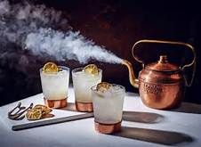 three glasses filled with ice and lemons next to a copper kettle