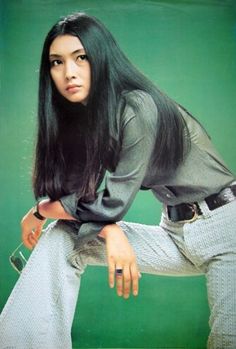 a woman with long black hair sitting on top of a green background wearing white pants