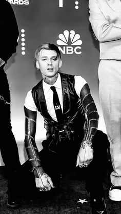 black and white photograph of a man dressed in punk clothing sitting on the floor with two people standing behind him