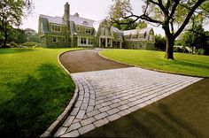 a brick driveway leading to a large house in the middle of a grassy area with trees