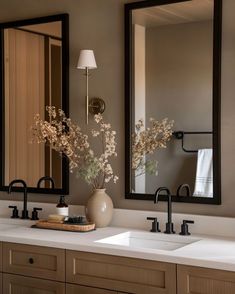 two sinks and mirrors in a bathroom with flowers on the counter top next to them