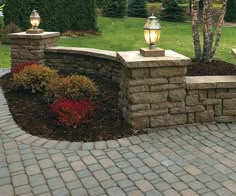 a stone wall with two lamps on top and some bushes in the back ground next to it