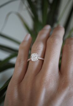 a woman's hand with a diamond ring on top of her finger, next to a potted plant