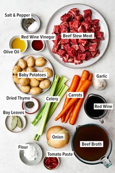 the ingredients for beef stew laid out on a white counter top, including carrots, celery, broth, garlic, red wine, and baby potatoes