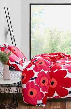 a bed with red flowers on it in front of a large window next to a small table