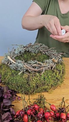 a woman is making a wreath out of moss and berries on a wooden table with purple flowers