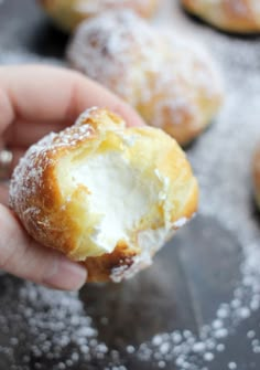 a hand holding a powdered sugar doughnut in front of other pastries on a baking sheet