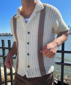 a man wearing a white crocheted cardigan standing next to a fence near the water