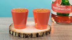 two shot glasses filled with watermelon sitting on top of a wooden coaster next to a bottle of watermelon