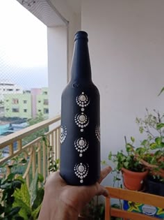 a hand holding up a black bottle with white designs on the outside and inside, in front of some potted plants