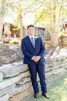 a man in a suit leaning against a stone wall with his hands on his hips