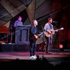 two men playing guitars on stage with microphones in the foreground and one man singing