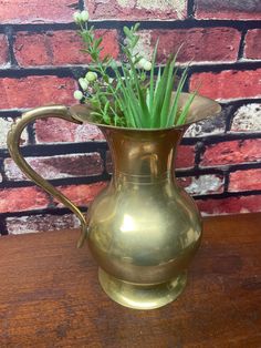 a brass pitcher with flowers in it on a wooden table next to a brick wall