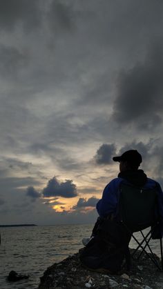 a man sitting in a chair on top of a rock near the ocean at sunset