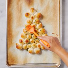 a person holding an orange brush over some food on a baking sheet with buns in the shape of a christmas tree