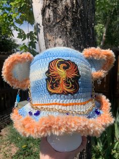 a hand holding up a crocheted hat with an orange and blue bear on it