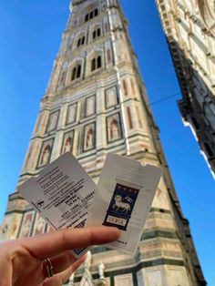 someone holding up two tickets in front of a tall building with a clock on it