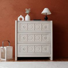 a white dresser sitting next to a lamp on top of a hard wood floor in front of a brown wall