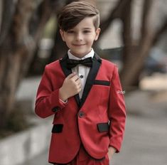 a young boy in a red suit and bow tie standing on the sidewalk with his hands in his pockets