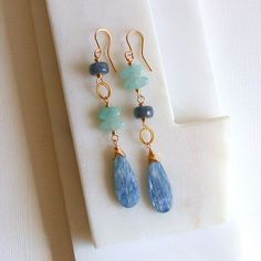 A pair of asymmetrical earrings featuring a large, polished drop of kyanite and topped with grey jade and aquamarine. earrings measure approximately 3 inches. Shown in 14k gold fill, also available in sterling silver.  I use only the finest quality materials to make my pieces. Each stone is carefully selected and I never use plated materials. All chains, wires and findings are sterling silver or 14k gold fill. I guarantee all of my work, including materials as well as the craftsmanship. I will b Gemstone Earring Ideas, Artisan Dangle Earrings With Gemstone Accents, Asymmetrical Beaded Earrings, Asymmetrical Earrings Handmade, Assymetrical Earrings Style, Handmade Blue Aquamarine Earrings, Kyanite Earrings, Natural Stone Earrings, Asymmetrical Earrings