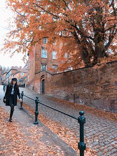 a woman walking down a sidewalk next to a tree with orange leaves on the ground