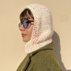 a woman wearing sunglasses and a knitted hoodie looks off into the distance while standing against a wall