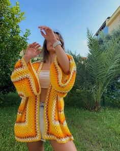 a woman standing in the grass wearing a yellow and white crochet cardigan