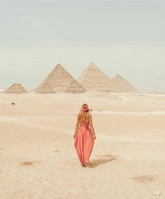 a woman in a red dress walking through the desert with three pyramids behind her