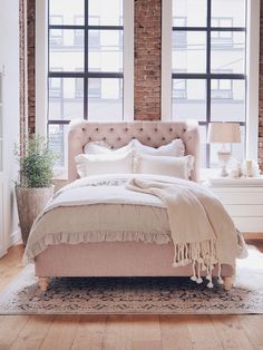 a bedroom with brick walls and large windows, a bed in the foreground is surrounded by white furniture