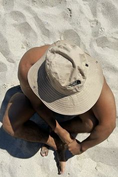 a man in a hat sitting on the beach looking down at his feet and back