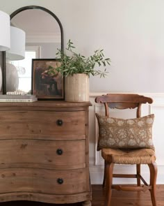 a wooden dresser sitting next to a lamp and a chair with a plant on top of it