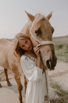 a woman in white dress standing next to a brown horse on dirt road near grass field