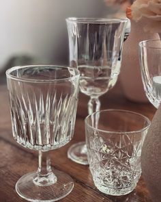 three clear wine glasses sitting on top of a wooden table next to a vase with flowers in it