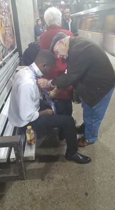 an old man getting his tie put on by another older gentleman at the train station