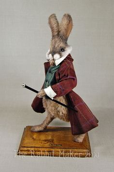 a stuffed rabbit dressed in a red coat and green scarf holding a cane on a wooden base