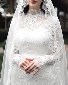 a woman wearing a white wedding gown and veil with her hands on her chest, standing outside