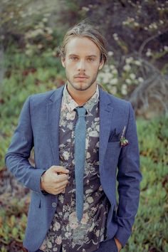 a man with long hair wearing a blue suit and flowered shirt is standing in the woods