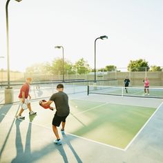 people playing tennis on an outdoor court with sun shining through the trees and buildings in the background