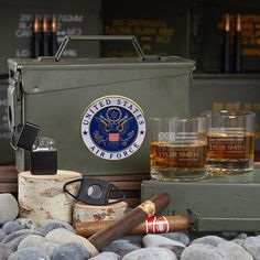 an army lunchbox, two glasses and cigars are sitting on the rocks next to each other