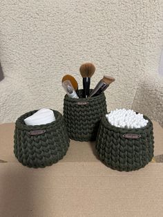 two green knitted baskets sitting on top of a cardboard box filled with makeup brushes