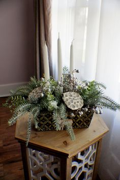 a wooden table topped with a vase filled with greenery and candles next to a window