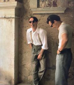 two men standing next to each other in front of a stone wall wearing suspenders