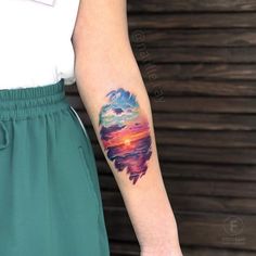 a woman's arm with a colorful sunset tattoo on her left forearm and an ocean wave coming out of the water