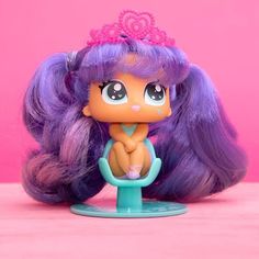 a doll with long purple hair and a tiara on her head sitting in front of a pink background