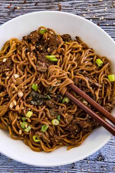 a white bowl filled with beef and noodles on top of a wooden table next to chopsticks